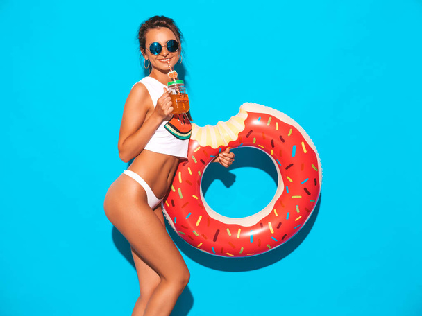 Young beautiful sexy smiling hipster woman in sunglasses.Girl in white summer underpants and topic with donut lilo inflatable mattress.Positive female going crazy.Posing near blue wall - Photo, Image