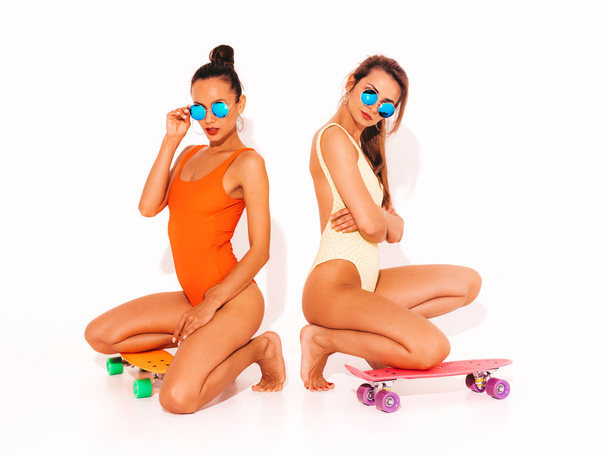 Two beautiful sexy smiling hipster women in summer colorful swimwear bathing suits.Trendy Girls in sunglasses. Positive models sitting on the floor with colorful penny skateboards.Isolated on white - 写真・画像