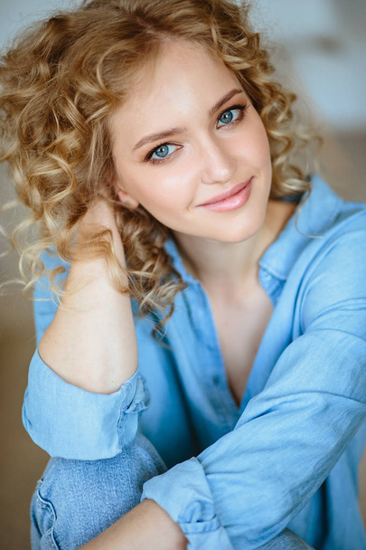 Pretty cheerful blonde young woman with curly hair and charming smile resting in her home, room with modern scandinavian interior - Fotografie, Obrázek