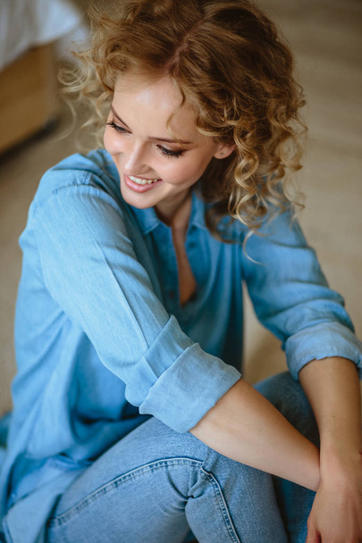 Pretty cheerful blonde young woman with curly hair and charming smile resting in her home, room with modern scandinavian interior - Foto, afbeelding