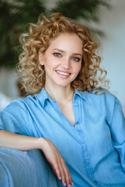 Pretty cheerful blonde young woman with curly hair and charming smile resting in her home, room with modern scandinavian interior - Foto, Bild