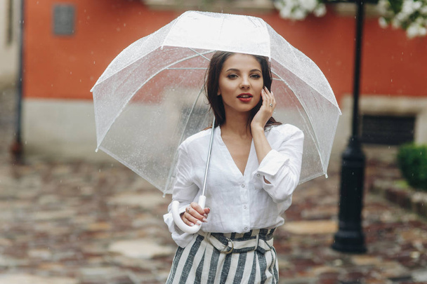 Beautiful girl dreamily walking around cozy city street with umbrella, casual style. It's summer rain. - 写真・画像