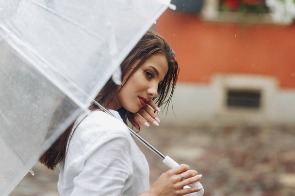 junge Frau posiert, während sie mit Regenschirm durch die Stadt läuft. Es regnet im Sommer. - Foto, Bild