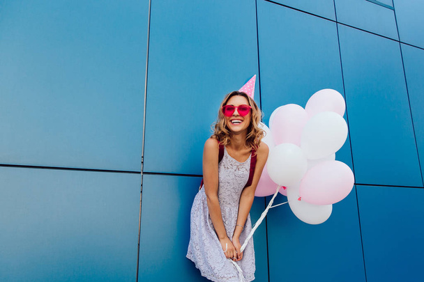 Happy woman in birthday hat standing with balloons near the urban wall - 写真・画像