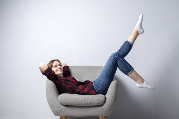 young girl is sitting on a soft comfortable chair against a white wall with her legs raised up, a hipster student is resting and dreaming, copy space - 写真・画像
