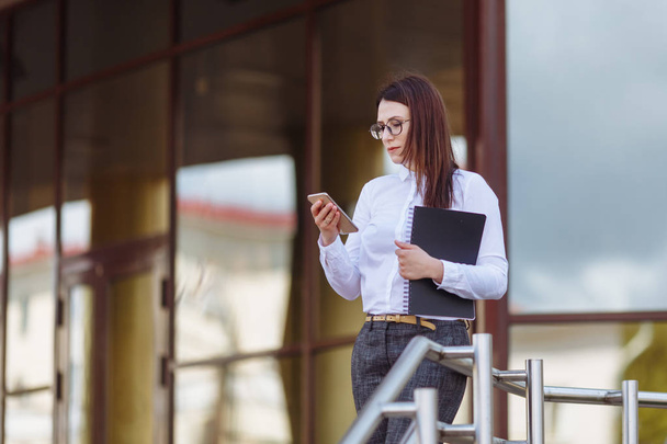 Portrait young business woman wearing white shirt using smartphone out doors. Female reading sms message in working process. - 写真・画像