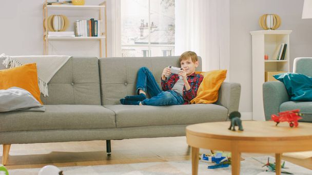 Очаровательный мальчик лежит на диване, играя в видеоигру на ТВ-консоли, используя джойстик-контроллер. Мальчик играет в настольную игру дома
. - Фото, изображение