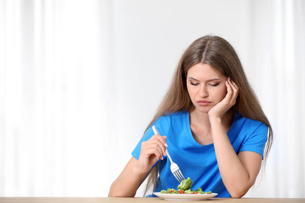 Portrait of unhappy woman eating broccoli salad at table on light background - Foto, Bild