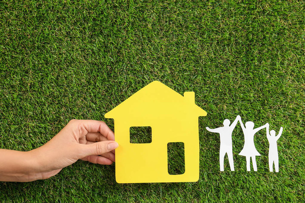 Woman holding yellow paper house near figures of happy family on green grass, top view - Photo, Image