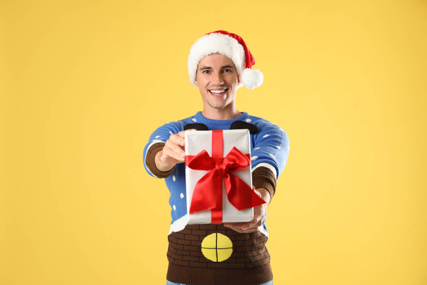 Happy man in Christmas sweater and Santa hat holding gift box on yellow background - Foto, Imagem