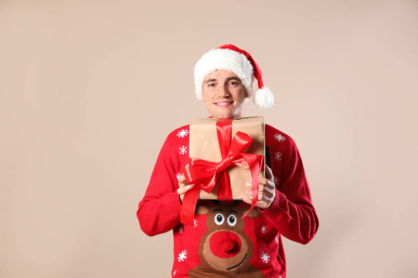 Happy man in Christmas sweater and Santa hat holding gift box on beige background - Фото, изображение