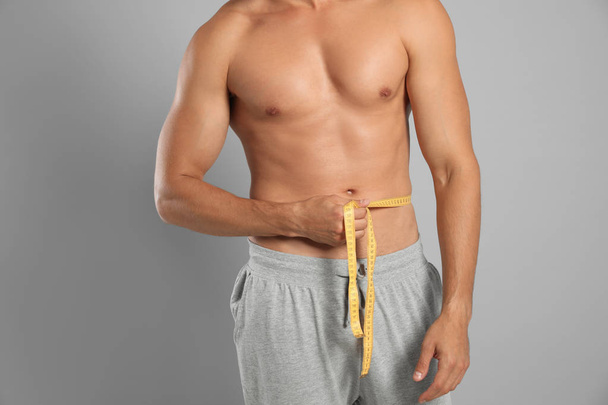 Young man with slim body using measuring tape on grey background, closeup view - Photo, image