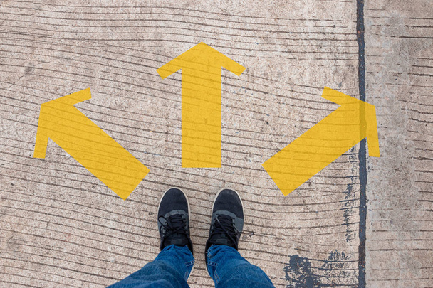 Persona che indossa scarpe da ginnastica in piedi su una strada di cemento con frecce gialle, concetto decisionale
 - Foto, immagini
