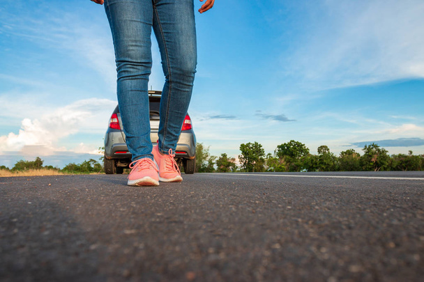 ジーンズを履いた女性、高速道路を歩くスニーカー昼間に駐車中の高速道路の車の上を歩く、明るい青空と雲の背景. - 写真・画像