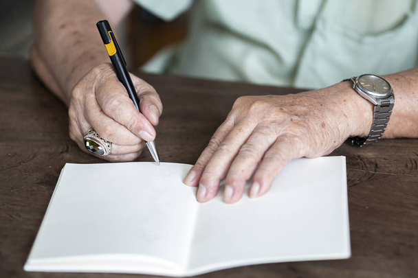 Senior man's hand schrijven brief op ivoor kleur papier, notebook, een zwarte mix zilveren pen, op houten achtergrond, close-up & macro shot, selectieve focus, briefpapier concept - Foto, afbeelding