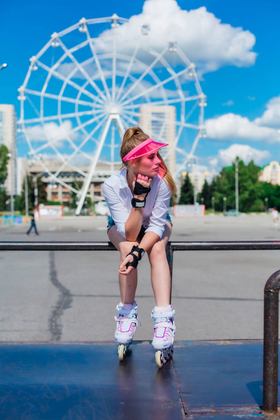 Portrait of an emotional girl in a pink cap visor wearing protective gloves and rollerblades sitting on the background of ferris wheel. - Photo, Image