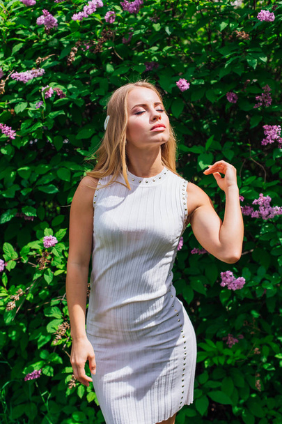 Portrait of a charming blond woman wearing beautiful white dress standing next to lilac bush. - 写真・画像
