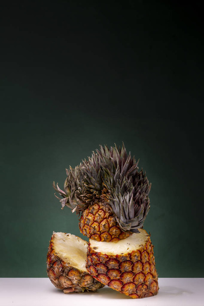 Upright standing sliced pineapple with yellow interior parts sticking out and extraordinary top on a white surface against a dark green background - Фото, изображение
