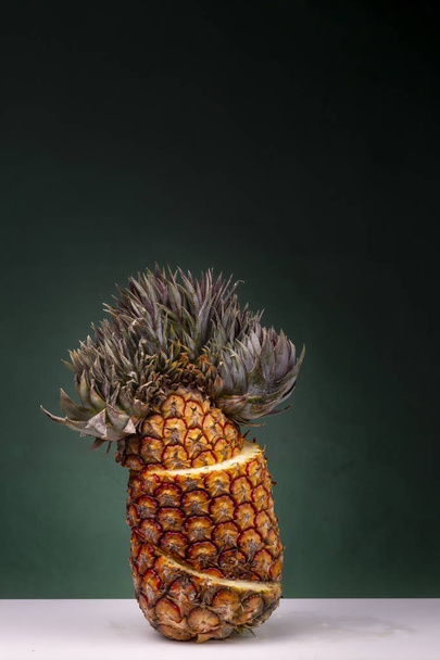 Upright standing sliced pineapple with yellow lines where you can see the yellow  pulp and extraordinary top on a white surface against a dark green background - Foto, Bild