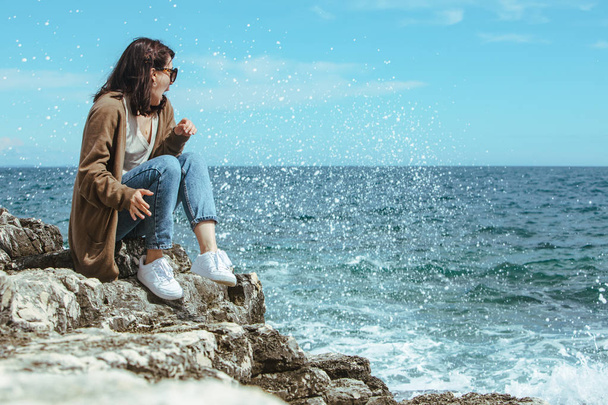 崖の上に座って海の景色を楽しむ女性。風の強い天気。晴れた日 - 写真・画像
