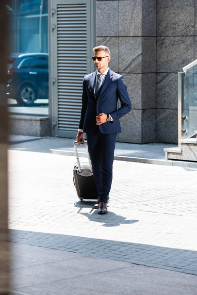 handsome man in suit and glasses holding suitcase and looking way  - Foto, afbeelding