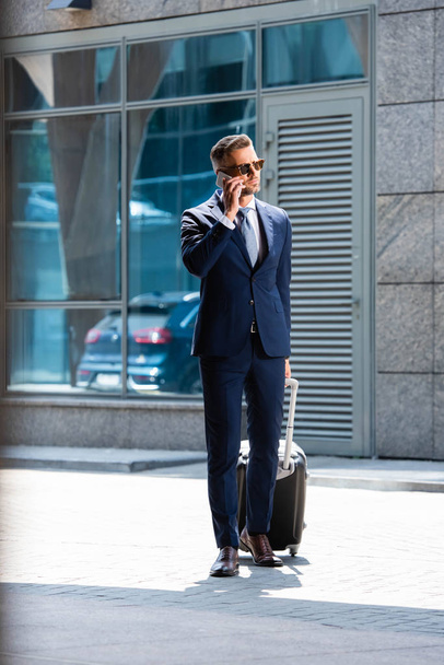 handsome man in suit and glasses holding suitcase and talking on smartphone  - Fotografie, Obrázek