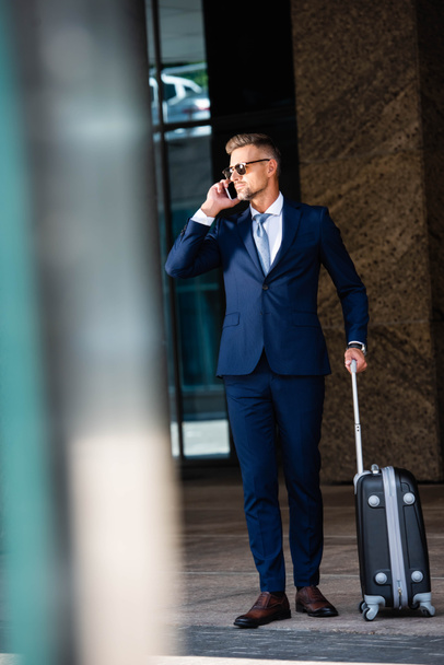 handsome businessman in suit and glasses talking on smartphone and holding suitcase  - Photo, Image