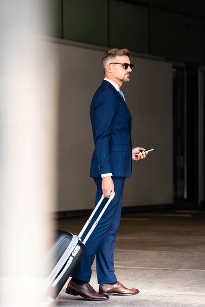 knappe zakenman in pak en glazen houden koffer en smartphone - Foto, afbeelding