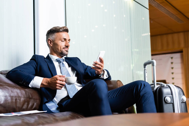 handsome businessman in suit using smartphone and holding cup  - 写真・画像