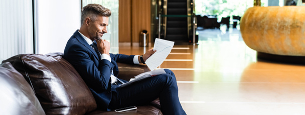 panoramic shot of handsome businessman in suit looking at papers  - Φωτογραφία, εικόνα