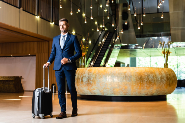 schöner Geschäftsmann im Anzug, der Koffer hält und wegschaut  - Foto, Bild