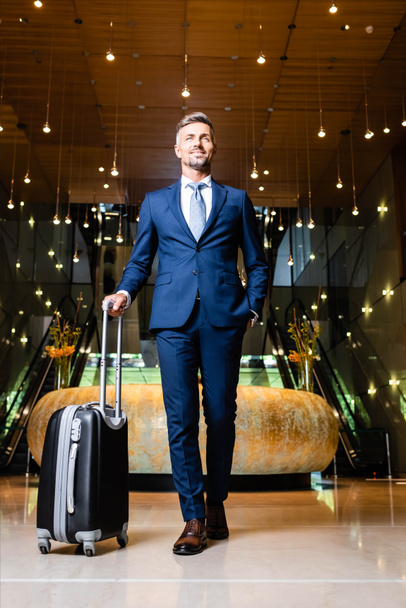 handsome businessman in suit with hand in pocket holding suitcase - Fotografie, Obrázek