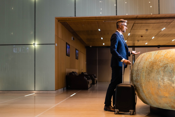 side view of handsome businessman in suit and glasses holding suitcase - Foto, imagen