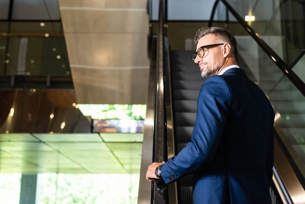 vista trasera de hombre de negocios guapo en traje y gafas en escalera mecánica
 - Foto, imagen