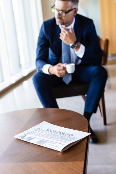 selective focus of newspaper on table and handsome businessman in suit with cup on background  - Zdjęcie, obraz