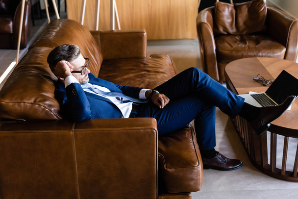 Вид сбоку красивого бизнесмена в костюме и очках, сидящего на диване
  - Фото, изображение