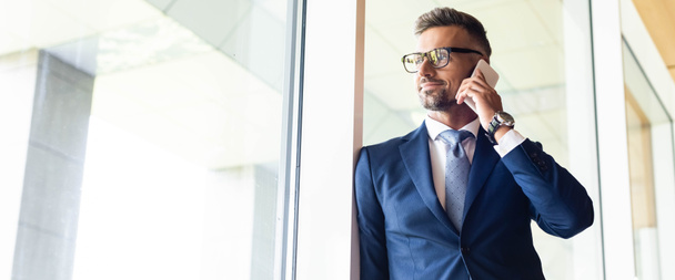 panoramic shot of handsome businessman in suit and glasses talking on smartphone  - Photo, Image