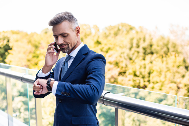 handsome businessman in formal wear talking on smartphone and looking at watch  - Foto, Imagem