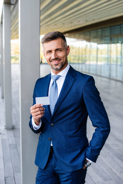 handsome businessman in formal wear with hand in pocket holding blank card  - Fotoğraf, Görsel