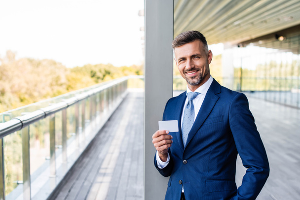handsome businessman in formal wear with hand in pocket holding blank card  - Фото, изображение