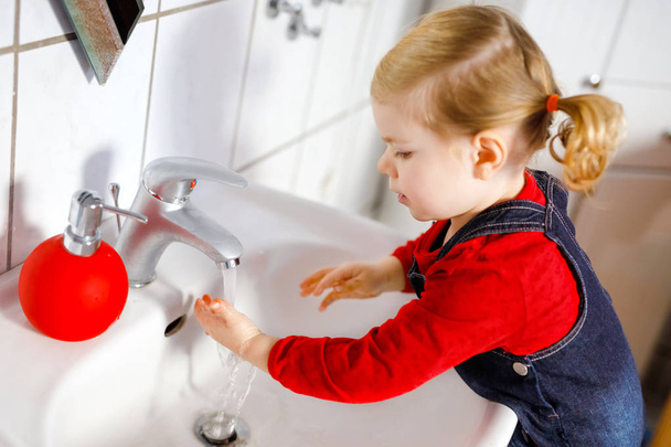 Χαριτωμένο κοριτσάκι που πλένει τα χέρια με σαπούνι και νερό στο μπάνιο. Αξιολάτρευτο παιδί μαθαίνει να καθαρίζει μέρη του σώματος. Πρωινή υγιεινή ρουτίνα. Ευτυχισμένο υγιές παιδί στο σπίτι ή στο νηπιαγωγείο. - Φωτογραφία, εικόνα