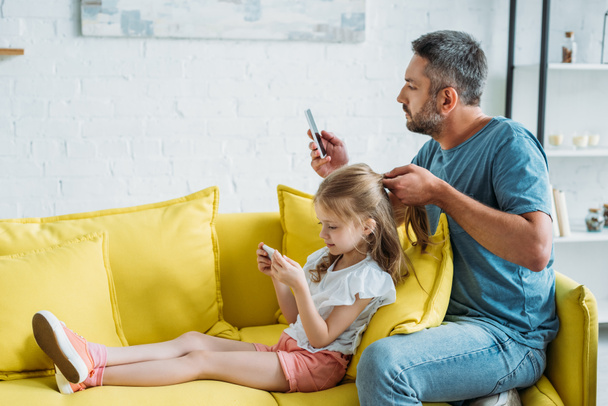 father touching daughters hair while sitting on couch and using smartphones together - Photo, Image