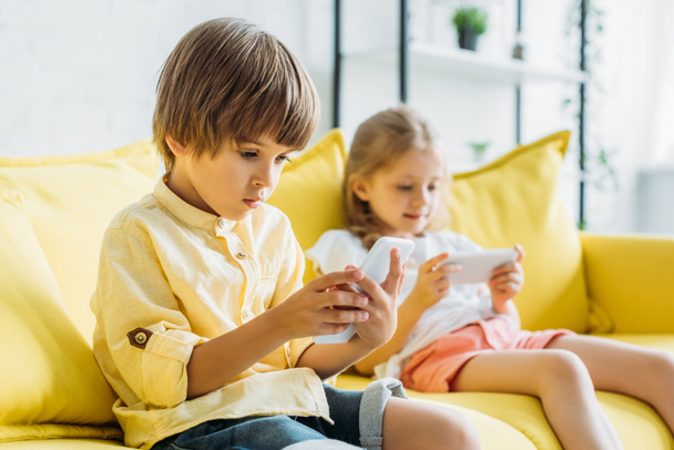 selective focus of cute boy with sister sitting on sofa and using smartphones at home - Φωτογραφία, εικόνα