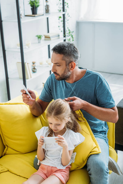 συγκεντρωμένη πατέρα αγγίζοντας τα μαλλιά της κόρης, ενώ κάθονται στον καναπέ και χρησιμοποιώντας smartphones μαζί  - Φωτογραφία, εικόνα