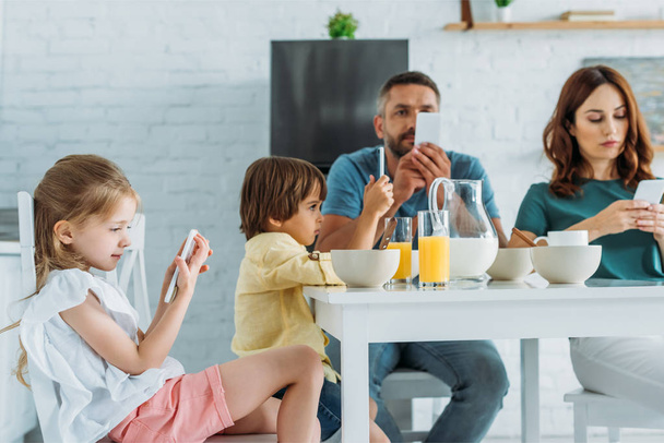 family using smartphones while sitting in kitchen near table with served breakfast - Foto, afbeelding