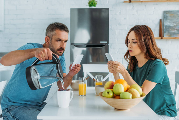 man with smartphone pouring water in cup while sitting in kitchen near wife using smartphone - Photo, Image