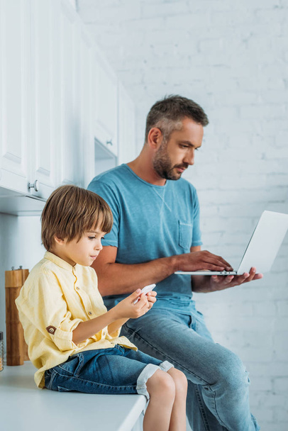 cute child using smartphome while sitting on kitchen counter near father using laptop - Фото, изображение