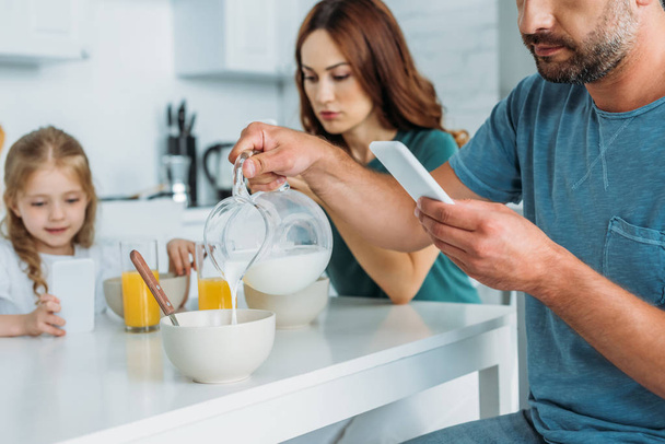 man with smartphone pouring milk in bowl while sitting at kitchen table near wife and daughter using smartphones - Фото, изображение
