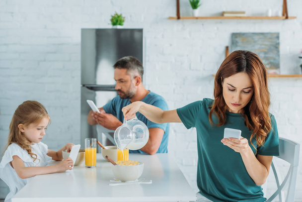woman with smartphone overfilling bowl with milk while sitting at kitchen table near husband and daughter using smartphones - Фото, изображение