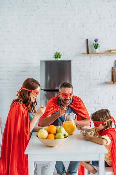 family in costumes of superheroes having breakfast in kitchen together  - Фото, изображение